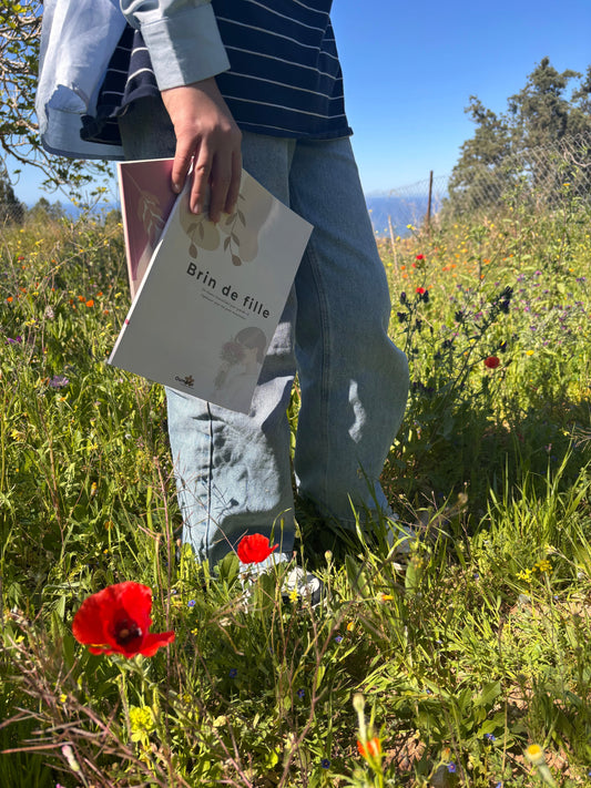 Adolescente tenant le livret « Brin de fille » dans un champ de fleurs sauvages, représentant la puberté, la douceur et l’accompagnement des jeunes filles musulmanes